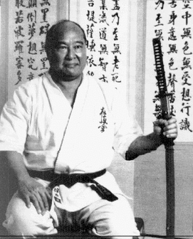 Mas Oyama kneeling in seiza holding a sheathed sword, seated before a wall of Japanese calligraphy.