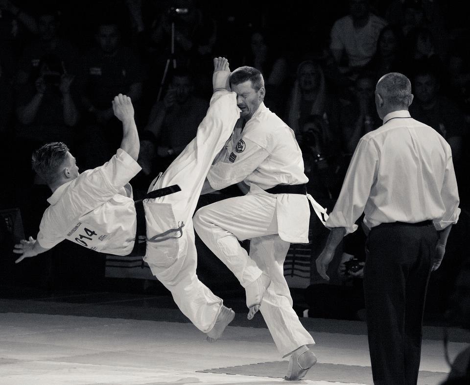 Two black-belt karateka in competition kumite, one landing a jumping kick as the referee watches.